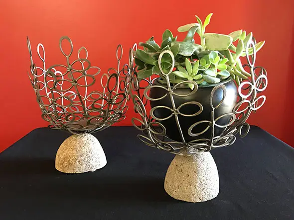 A flower pot of small green-leaf plants set in a designed metal cradle on top of decorative stone