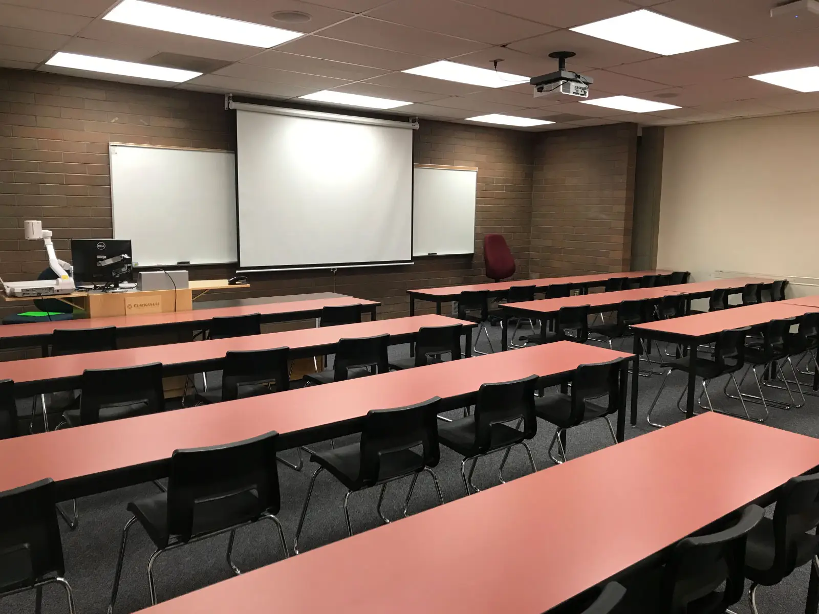 Rows of red tables and a podium in an Oregon CIty classroom with a projector screen against a brick wall