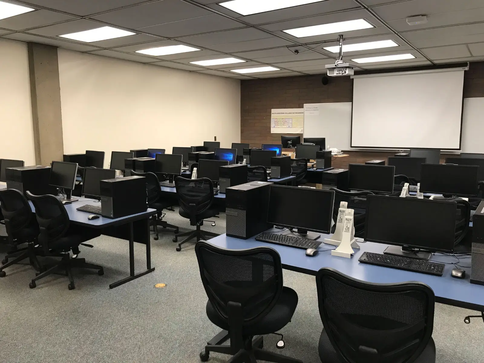Rows of tables with computers and comfortable, ergonomic chairs in front of a projector screen in a computer lab on the Oregon City campus