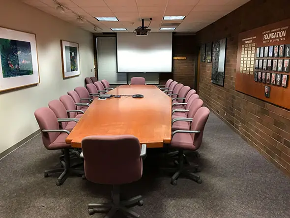 A long, skinny table with many chairs in front of a projector, surrounded by paintings and awards, in a conference room at the Oregon City campus