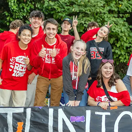 group of high school students pose, including student giving two thumbs up, student with red and white face paint and students giving a peace sign and bunny ears