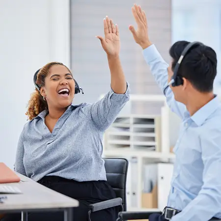 Two office workers in a corporate setting wearing headsets smile and high five.