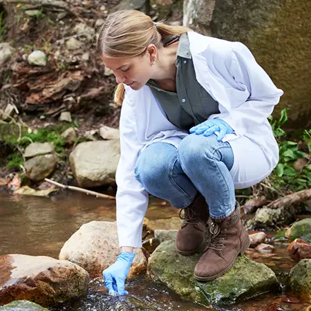Geology student with a white lab coat bending over to gather a water sample from a river.