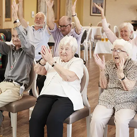 Group of older adults at a living facility sitting in chairs and raising their arms.