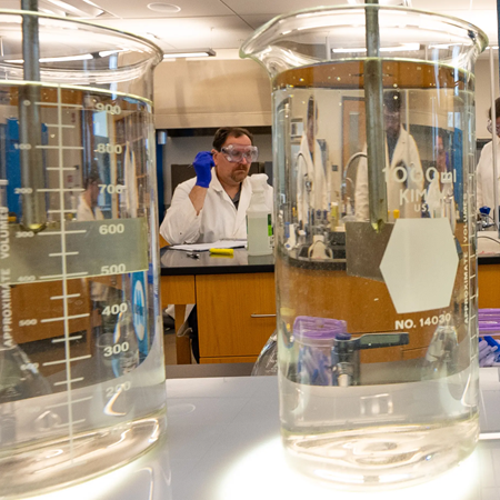 Chemistry lab: Students in white lab coats and protective eyewear can be seen through beakers of clear liquid in the foreground.