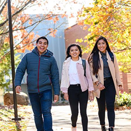 Three students smiling and walking on campus in front of campus building and fall foliage